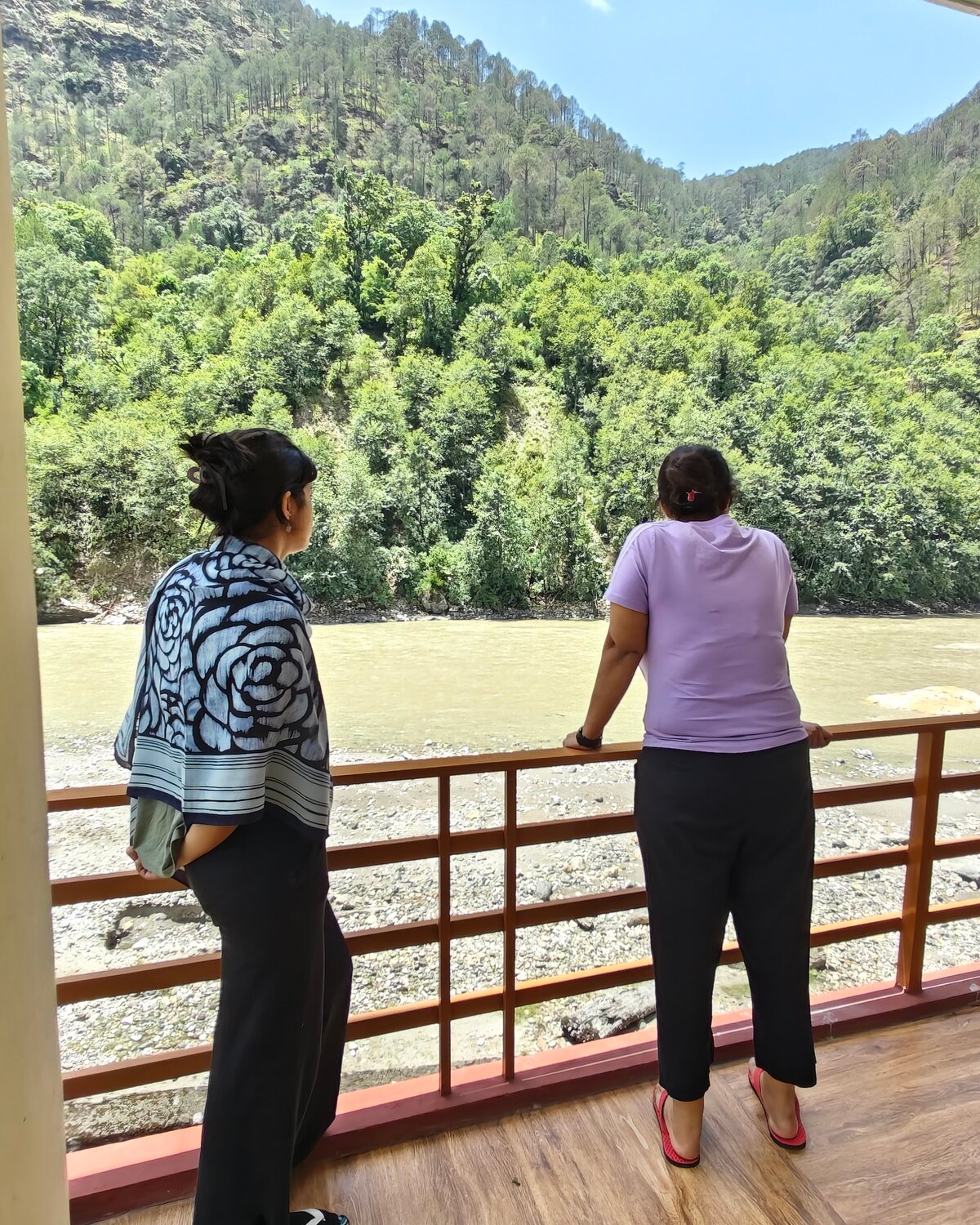 Two women looking out over the Bhagirathi River and forested mountains from a balcony railing